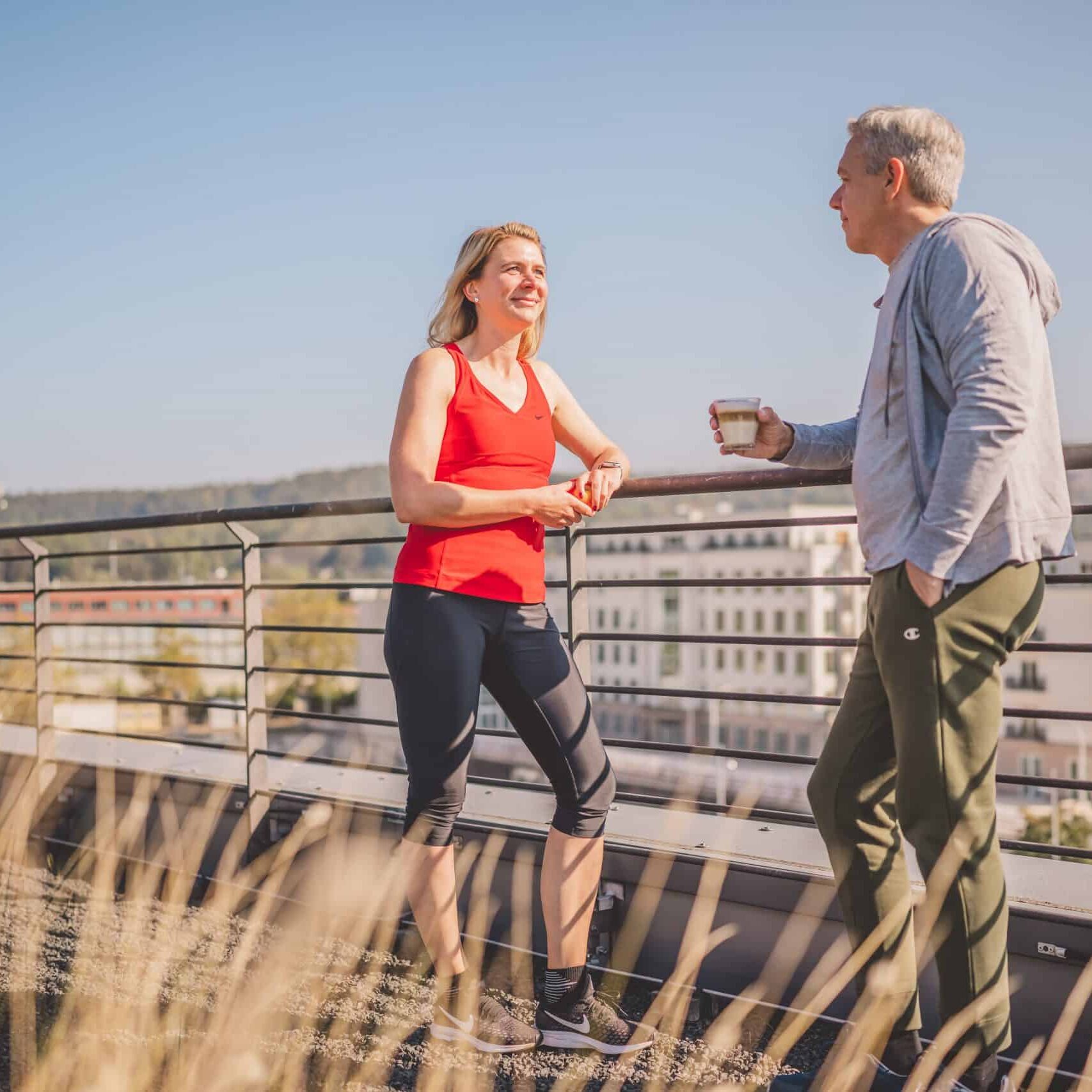 Zwei Menschen stehen auf einer Dachterrasse, der Mann hält einen Kaffee in der Hand, die Frau schaut ihn an