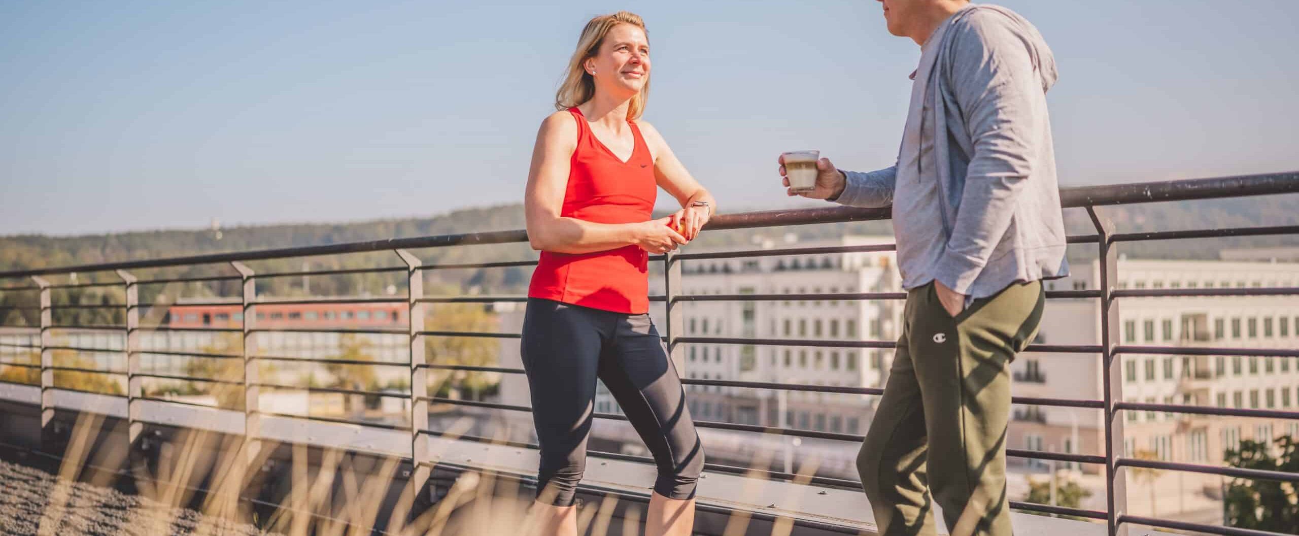 Zwei Menschen stehen auf einer Dachterrasse, der Mann hält einen Kaffee in der Hand, die Frau schaut ihn an