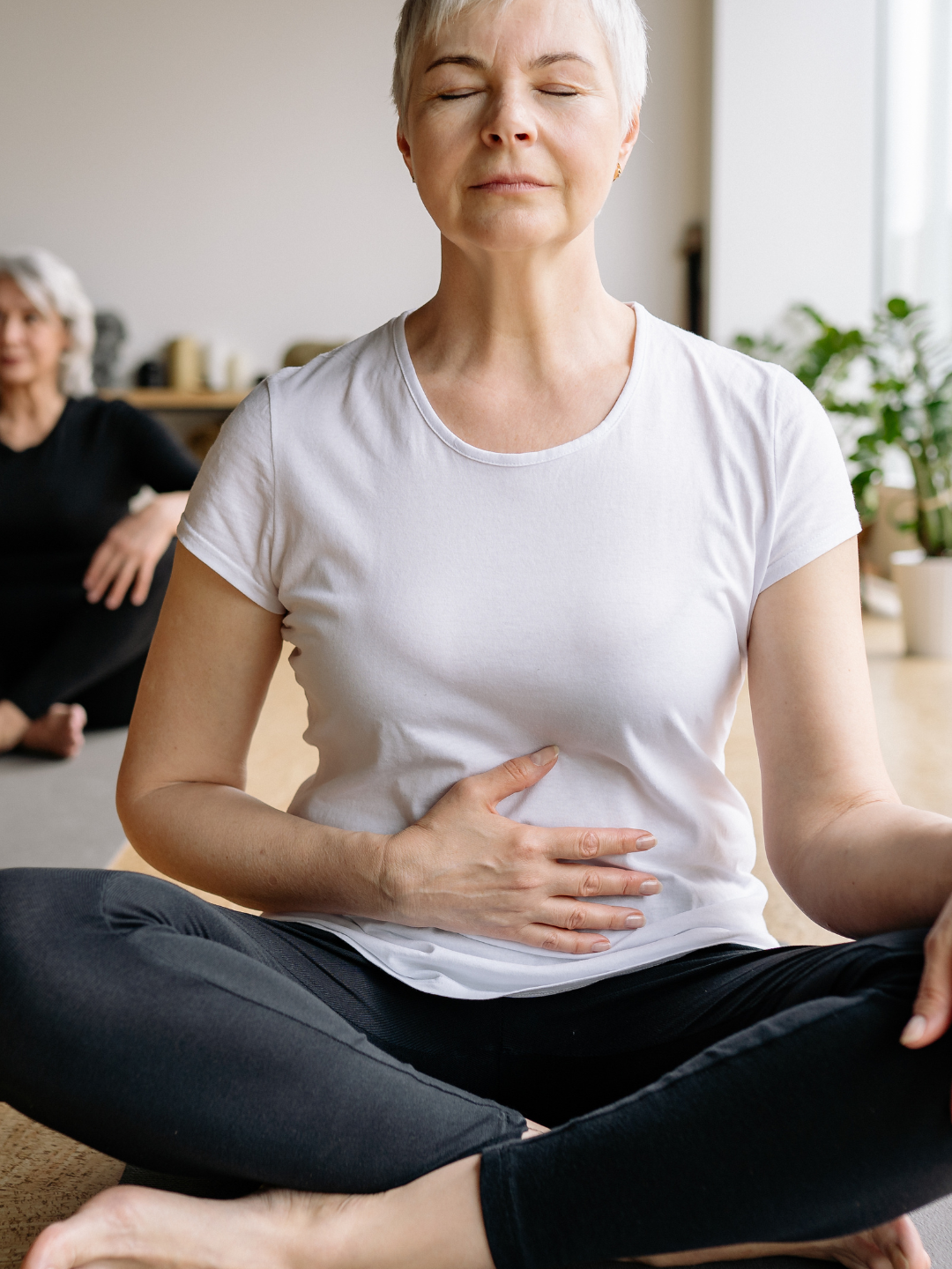 Ältere Frau sitzt im Schneidersitz auf einer Yogamatte mit geschlossenen Augen und berührt ihren Bauch.