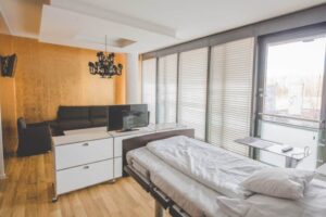 Modernes Zimmer mit Bett, Sofa, TV, schwarzem Kronleuchter und großen Fenstern mit Jalousien.