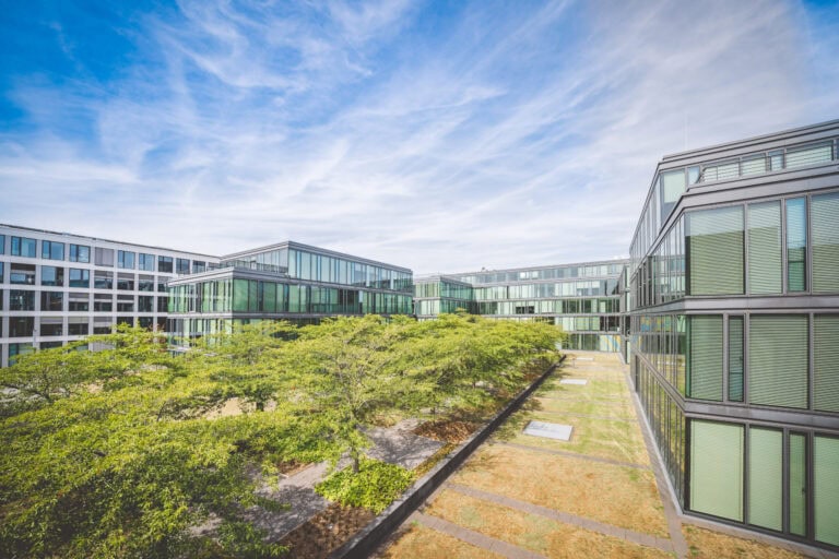 Moderne Bürogebäude mit Glasfassaden umgeben einen begrünten Innenhof unter blauem Himmel.