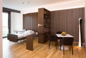 Helles Patientenzimmer mit Bett, Sitzecke und Holzmöbeln.