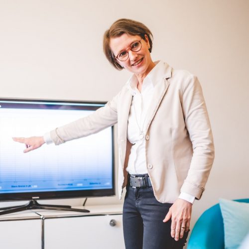 Nahaufnahme von Biofeedbacktherapeutin Britta Wiemann wie sie auf einen Bildschirm zeigt und lächelnd in die Kamera guckt.
